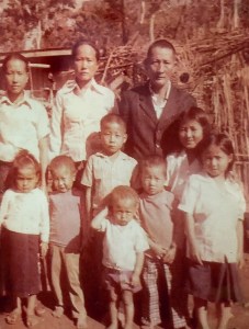 Author’s maternal grandmother and her mother as a child stand with their family members. The three adults are standing in the back. The children are standing in the front. They are all facing forward. The sun is shining. Behind them is a makeshift shelter and a wooden gate.