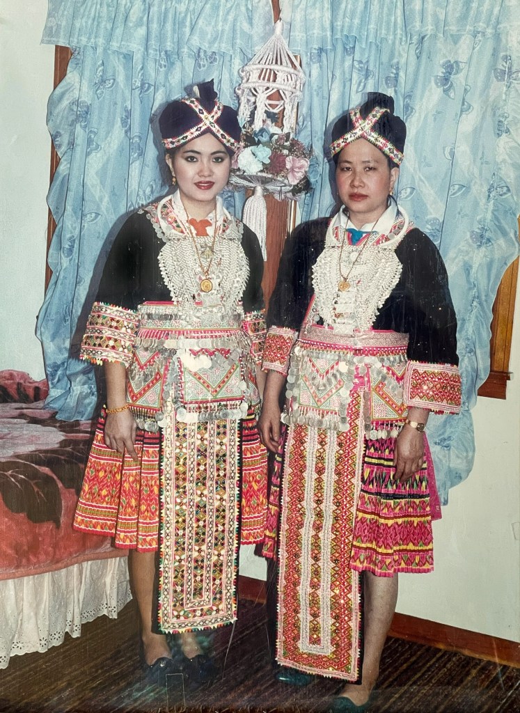 The author’s maternal grandmother and mother stand next to each other, dressed in traditional Hmong clothing. They are wearing a turban hat, an embroidered shirt, and a long pleated skirt. They accessorize with a silver necklace, a coin belt, and coin purses.
