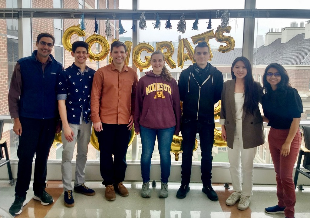 The author and her lab member are standing next to a microscope. They are wearing transparent goggles and white lab coats. They are smiling at the camera.