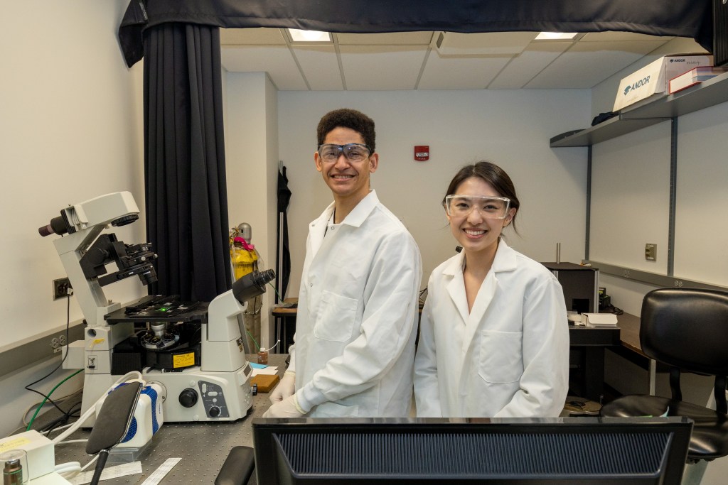 The author and her lab member are standing next to a microscope. They are wearing transparent goggles and white lab coats. They are smiling at the camera.