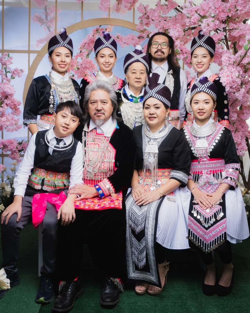 The author and her family members pose in a photography studio. They are wearing Hmong clothes. There are pink floral trees around and behind them.