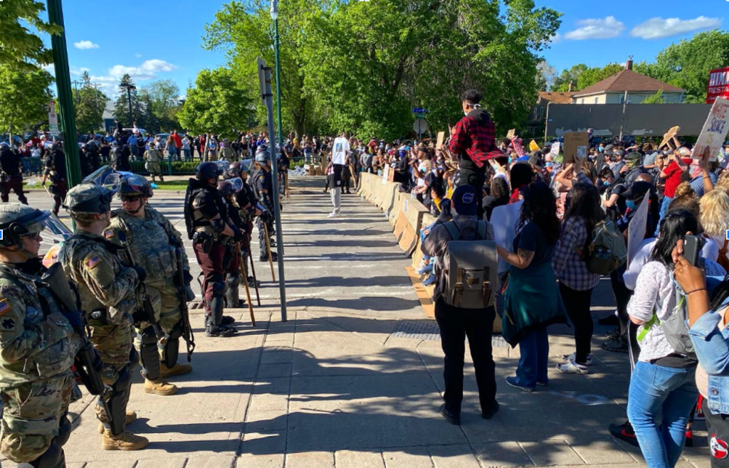 On the left side of the photo there is a line of national guard troops, in full gear, helmets, and with batons. There is some space between them, and a large crowd of protestors standing behind jersey barriers.