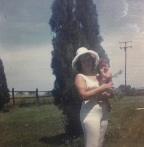 A polaroid photo of a Native American woman wearing a white dress and white hat holding a baby in a grassy field with a gate, telephone poles, and pine trees in the background. The woman is the author’s grandmother and the baby is the author’s father