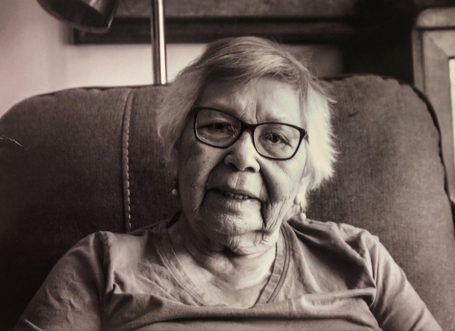 Black and white portrait of an elderly woman with short gray hair and glasses, sitting comfortably in a chair and smiling softly.
