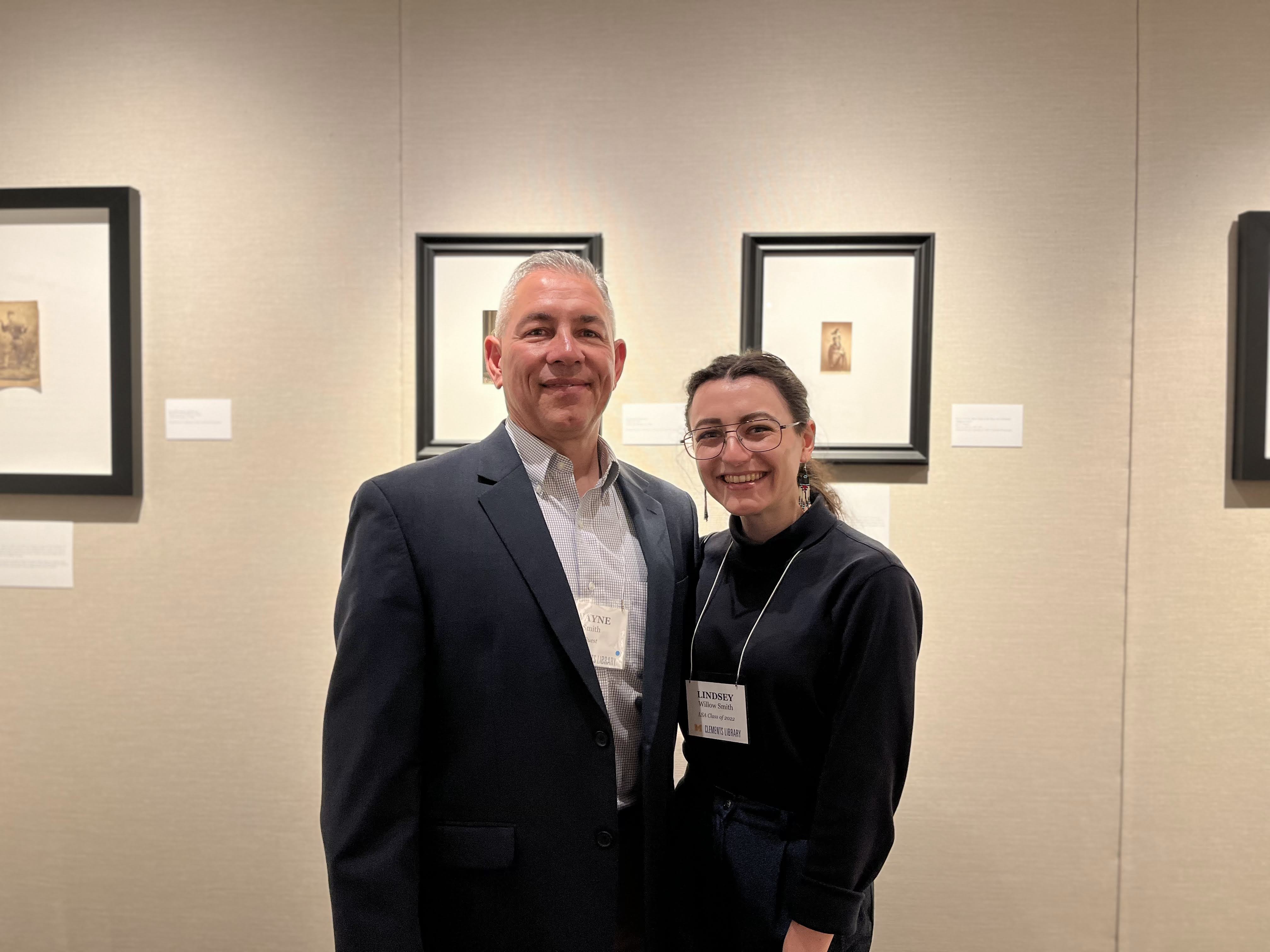 Two individuals posing for a photo in a gallery setting with framed artworks on the wall behind them.