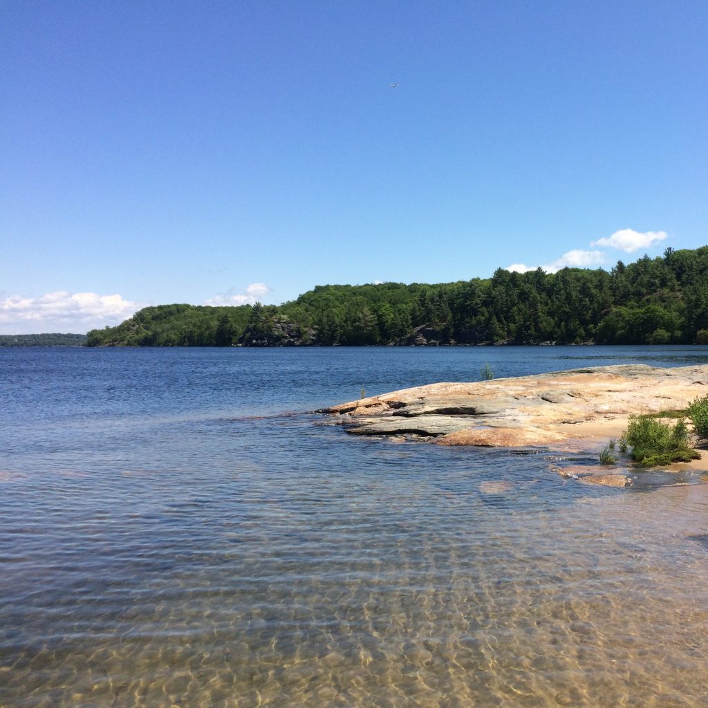 Summer on the Rez: a landscape photo of a sandy beach in the foreground next to a rock sloping into the water. The background is blue water and rocky shoreline covered in pines and deciduous trees.