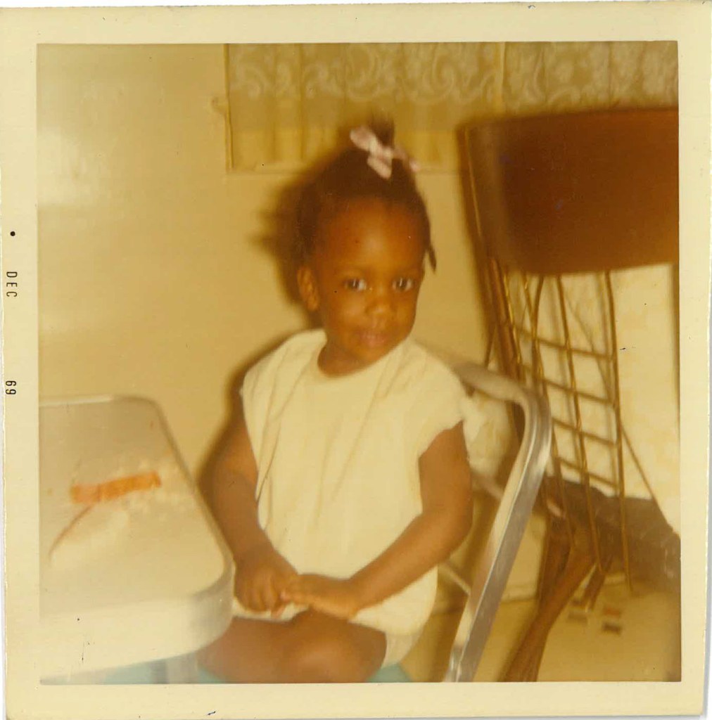 Faded photograph of Toddler Shari at home sitting at the table, 1969.