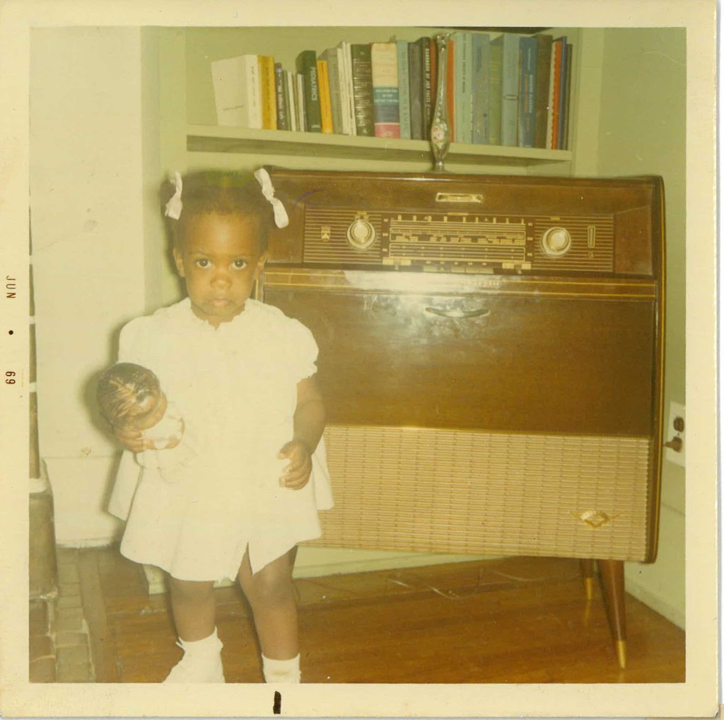 Faded, square photograph of toddler Shari at home holding a dolly, 1969.